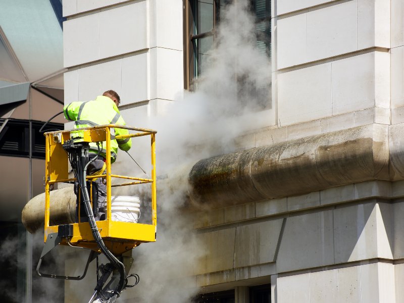 professional in yellow jacket and on lift cleaning the siding of building
