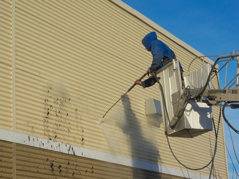 professional in blue hoodie washing siding of brown building